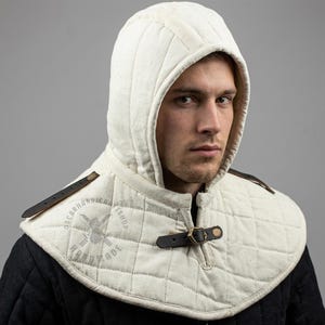 May include: Cream-colored quilted hood with a diamond pattern and a leather strap closure. The hood is part of a medieval-style garment, featuring a padded design for protection. The garment has the text "OSCARHANDICRAFTSHOP HAND MADE" printed on it.