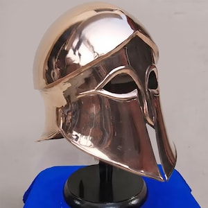 May include: A bronze-coloured helmet with a polished finish, featuring eye openings and cheek guards. The helmet is displayed on a black stand, highlighting its reflective surface and historical aesthetic. The design suggests a historical or fantasy theme.