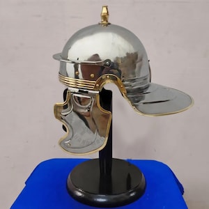 May include: A polished silver Roman helmet with gold-colored accents, including a brim and cheek guards. The helmet is displayed on a black stand with a blue surface in the background. The text "HANDMADE" is visible on the cheek guard.