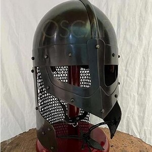 May include: A black metal Viking helmet with a riveted construction and a face guard. The helmet features a mesh visor and is displayed on a red stand. The helmet is a replica of historical armor.