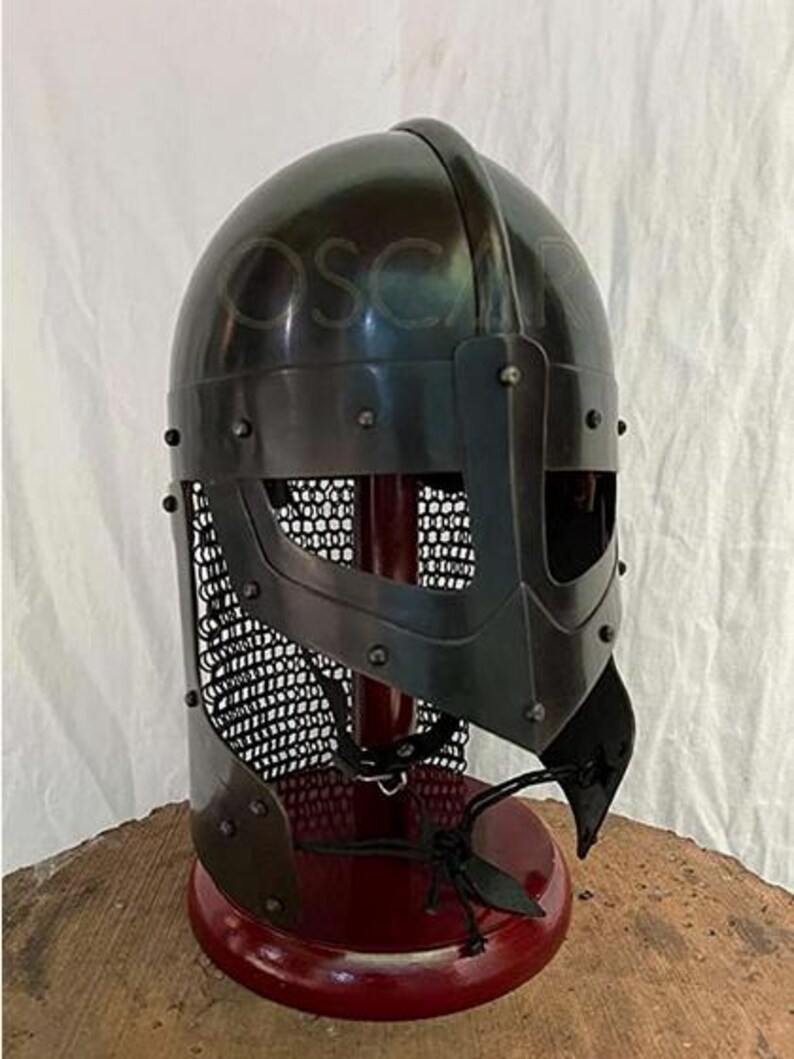 May include: A black metal Viking helmet with a riveted construction and a face guard. The helmet features a mesh visor and is displayed on a red stand. The helmet is a replica of historical armor.