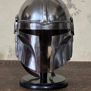 May include: A metallic silver Mandalorian helmet, displayed on a black stand. The helmet has a distinctive T-shaped visor and a sleek, polished finish. The image is well-lit, highlighting the helmet's details and contours. The helmet is a replica of the one worn by the Mandalorian.