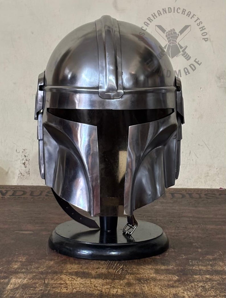 May include: A metallic silver Mandalorian helmet, displayed on a black stand. The helmet has a distinctive T-shaped visor and a sleek, polished finish. The image is well-lit, highlighting the helmet's details and contours. The helmet is a replica of the one worn by the Mandalorian.