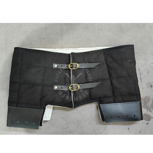 Könnte beinhalten: Schwarze gepolsterte Rüstung mit Lederdetails und Messingschnallen. Die Rüstung hat ein gestepptes Design und wird mit zwei Lederriemen befestigt. Das Produkt ist auf Schutz und Haltbarkeit ausgelegt.