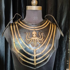 Könnte beinhalten: Eine schwarze und goldfarbene Brustplatte aus Metall mit einem Oktopus-Design. Die Brustplatte hat horizontale goldene Bänder und Nieten. Der Oktopus ist auf der Brustplatte zentriert. Die Brustplatte wird auf einer Schaufensterpuppe ausgestellt.