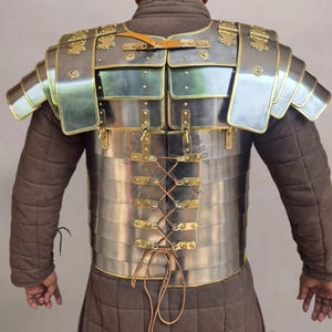 May include: A silver-colored Roman lorica segmentata armor with brass accents and leather straps. The armor is composed of overlapping metal plates and features shoulder guards. The back of the armor has a laced closure. The armor is worn over a brown padded garment.