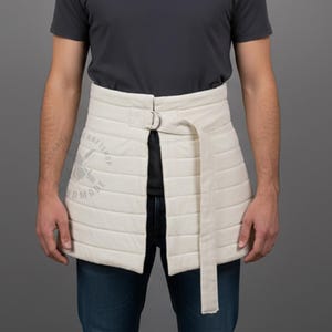 Puede incluir: Delantal acolchado color crema con diseño envolvente. El delantal presenta costuras horizontales, un cierre de hebilla metálica y un cinturón largo. El delantal se lleva sobre unos vaqueros oscuros y una camiseta gris. El delantal tiene el texto "CRAFTSHOP" y "DMADE".