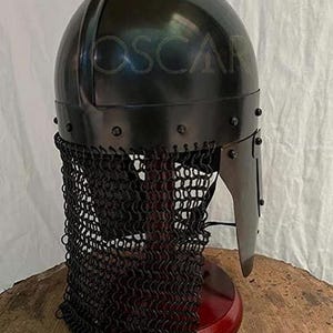 May include: A black medieval helmet with a chainmail neck guard. The helmet has a rounded top and a visor. The word "OSCAR" is faintly visible on the helmet. The helmet is displayed on a wooden stand.