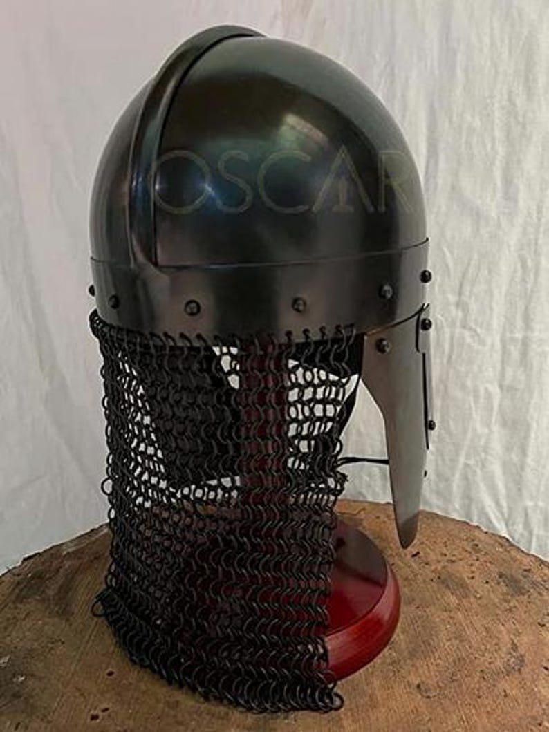 May include: A black medieval helmet with a chainmail neck guard. The helmet has a rounded top and a visor. The word "OSCAR" is faintly visible on the helmet. The helmet is displayed on a wooden stand.