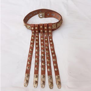 Puede incluir: Un cinturón de cuero marrón con detalles metálicos decorativos y seis correas de cuero colgantes. Cada correa está adornada con tachuelas metálicas y extremos metálicos decorativos. El cinturón presenta una hebilla metálica y cuadrados metálicos decorativos.