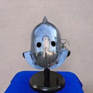 Puede incluir: Un casco pulido de color plateado con una parte superior puntiaguda y agujeros para los ojos. El casco tiene una visera curva y se exhibe sobre un soporte negro con una base redonda. El fondo es de color beige claro.