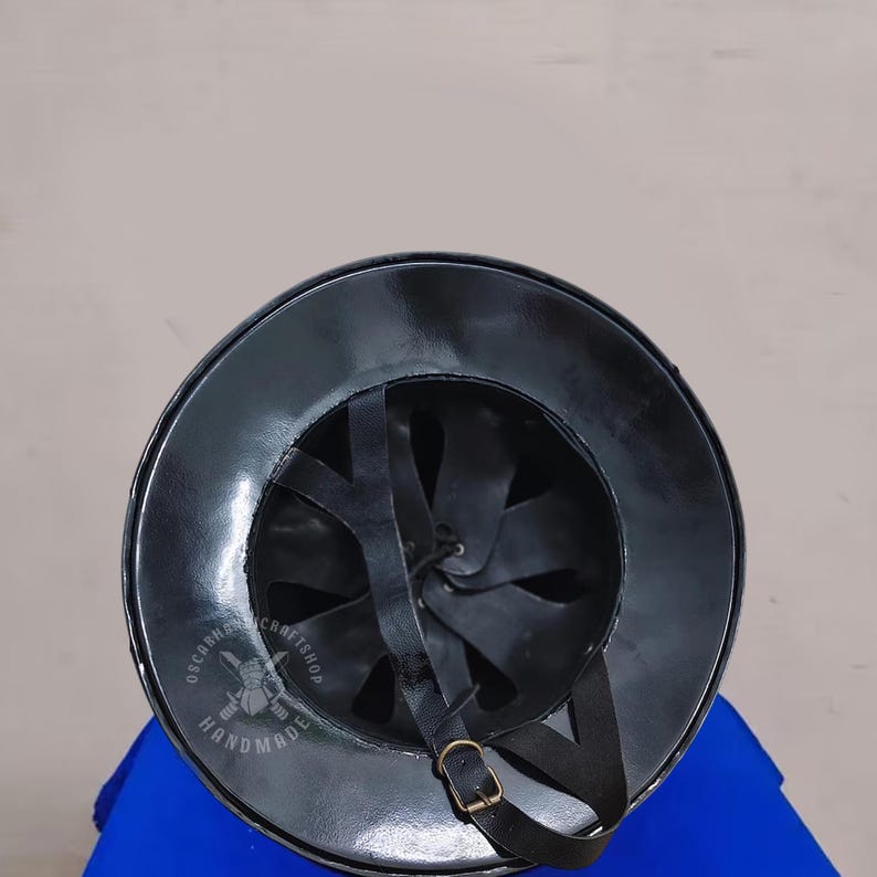 May include: A black, handcrafted helmet with a wide brim and leather straps. The interior features a series of leather straps for a secure fit. The helmet has a shiny, reflective surface and a brass buckle. The product is labeled "OSCARNA CRAFTSHOP HANDMADE".