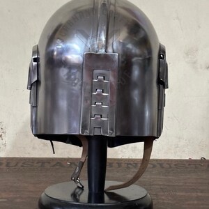May include: A metallic silver helmet with a rounded top and rectangular side panels. The helmet is displayed on a black stand with a leather strap. The helmet has a series of rectangular accents on the back.