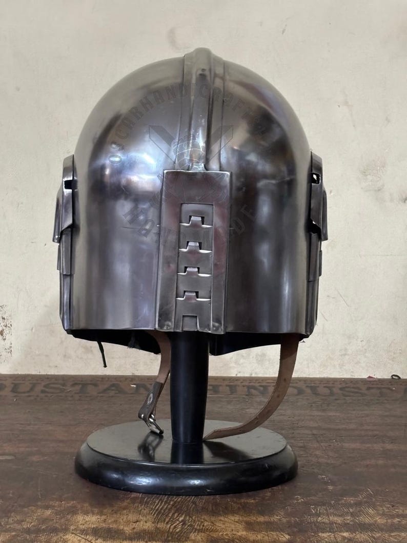May include: A metallic silver helmet with a rounded top and rectangular side panels. The helmet is displayed on a black stand with a leather strap. The helmet has a series of rectangular accents on the back.