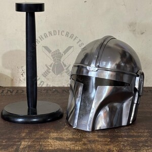May include: A polished metal helmet with a visor, next to a black helmet stand. The helmet has a distinctive shape with angular features. The stand is round at the base and top, with a vertical post.