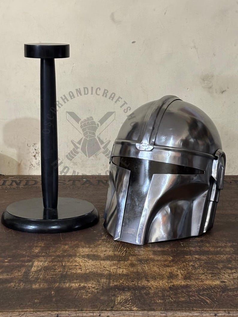 May include: A polished metal helmet with a visor, next to a black helmet stand. The helmet has a distinctive shape with angular features. The stand is round at the base and top, with a vertical post.