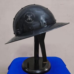 May include: A black metal helmet with a wide brim, displayed on a black stand. The helmet has a rounded top and is riveted along the edges. The text "OSCARHANDICRAFTSHOP HANDMADE" is visible on the helmet.
