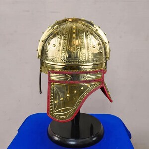 May include: A gold-colored helmet with intricate embossed patterns and small, round studs. The helmet features a red leather neck guard with decorative stitching. It is displayed on a black stand against a blue background.