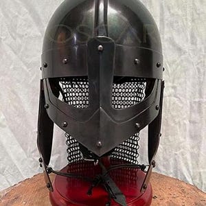May include: A black metal Viking helmet with a rounded top, a face guard, and a chainmail neck protector. The helmet is mounted on a red wooden stand. The helmet is a replica of a historical artifact.