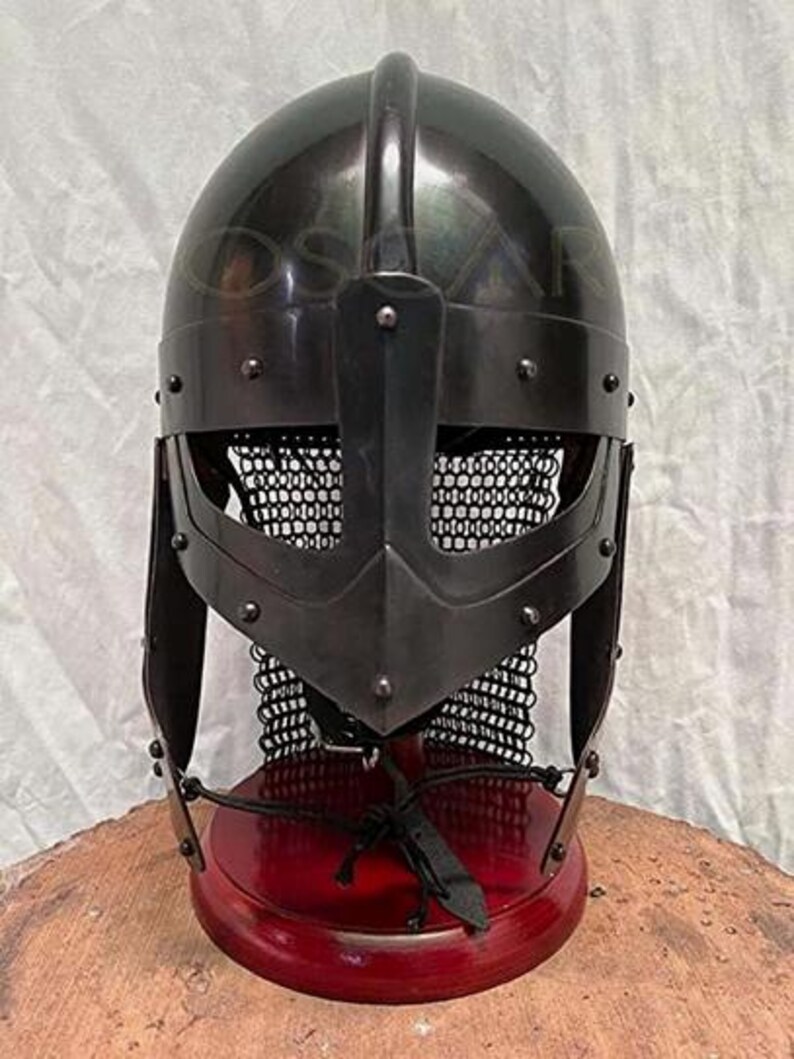 May include: A black metal Viking helmet with a rounded top, a face guard, and a chainmail neck protector. The helmet is mounted on a red wooden stand. The helmet is a replica of a historical artifact.