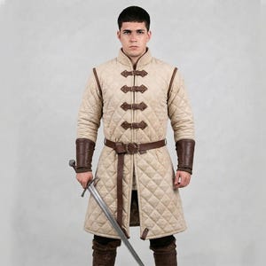 May include: A beige quilted gambeson jacket featuring a high collar, long sleeves, and a front closure with four brown leather straps and buckles. The garment includes a matching brown leather belt at the waist and dark brown leather bracers.