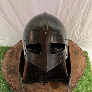 May include: A dark gray Viking helmet with a face guard and chainmail neck protection. The helmet is displayed on a weathered, round wooden base, set against a white backdrop and green grass.