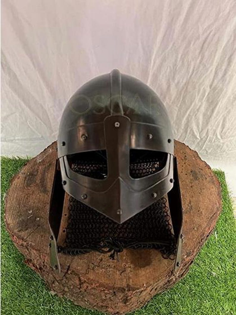 May include: A dark gray Viking helmet with a face guard and chainmail neck protection. The helmet is displayed on a weathered, round wooden base, set against a white backdrop and green grass.