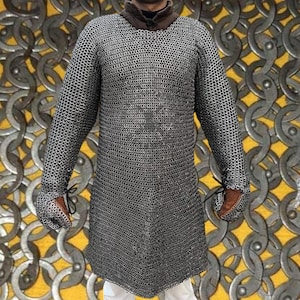 May include: A silver chainmail shirt with long sleeves. The shirt has a round neckline and reaches mid-thigh. The cuffs are finished with brown leather and black ties. The background features a pattern of interlocking metal rings.