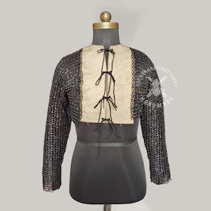 Könnte beinhalten: Ein mittelalterliches Kettenhemd mit langen Ärmeln, auf einer Schaufensterpuppe präsentiert. Das Hemd besteht aus kleinen, ineinandergreifenden Metallringen. Es hat eine zentrale beige Platte mit schwarzer Schnürung. Das Kleidungsstück ist für Kostümzwecke bestimmt.