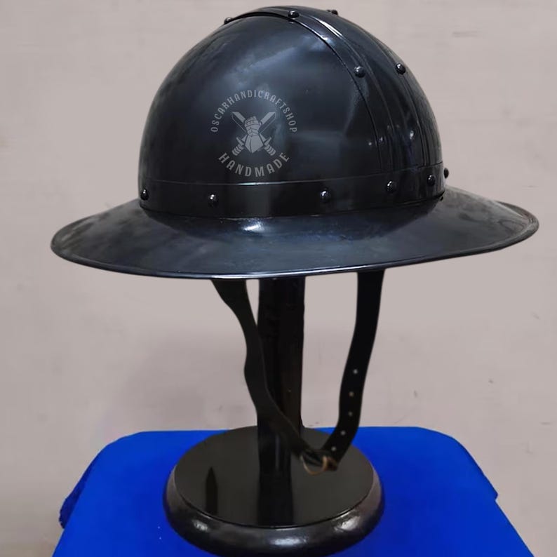 May include: A black metal helmet with a wide brim, riveted construction, and a leather chin strap. The helmet is displayed on a black stand with a round base. The text "OSCARHANDICRAFTSHOP HANDMADE" is visible on the helmet.
