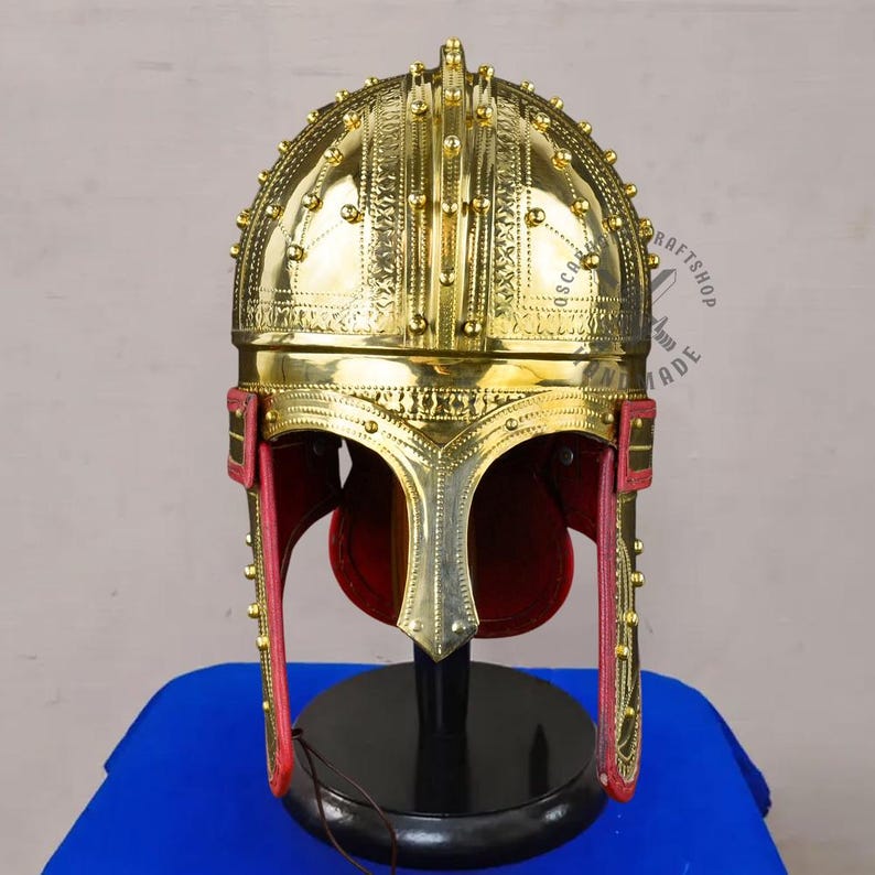 May include: A gold-colored helmet with intricate detailing, including small studs and decorative patterns. The helmet features a prominent face guard and red leather cheek pieces. It is displayed on a black stand against a blue background.