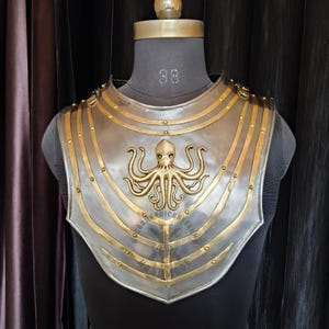 Può includere: Un'armatura pettorale color argento con dettagli dorati e un emblema di polpo. L'armatura è esposta su un manichino e presenta dettagli rivettati ed elementi di design curvi. Il testo "HANDMADE" è visibile.