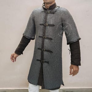 May include: A silver chainmail shirt with short sleeves and a leather-strapped front closure. The shirt has a long, tunic-like design and is worn over padded sleeves. The overall aesthetic is reminiscent of medieval armor.