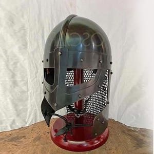 May include: A dark gray metal Viking helmet with a riveted construction. The helmet features a face guard and a mesh visor. The helmet is displayed on a red stand with a wooden base. The text "020" is visible on the helmet.