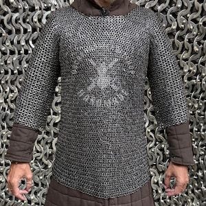 Puede incluir: Una camisa de cota de malla plateada con mangas largas y puños acolchados marrones. La camisa tiene un cuello redondo y una sección acolchada marrón en la parte inferior. La cota de malla está hecha de pequeños anillos de metal entrelazados.