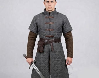 May include: A grey quilted gambeson with short sleeves, featuring a high collar and four brown leather buckle straps down the front. A brown leather belt with an attached pouch is worn at the waist over the garment, which is worn over brown sleeves.