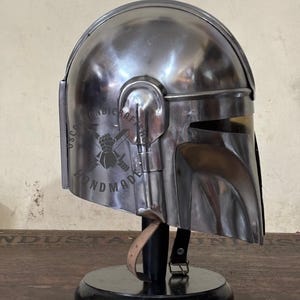 May include: A silver-colored helmet with a visor and ear pieces, displayed on a black stand. The helmet has a reflective surface and appears to be made of metal. The text "HANDMADE" is visible on the side.