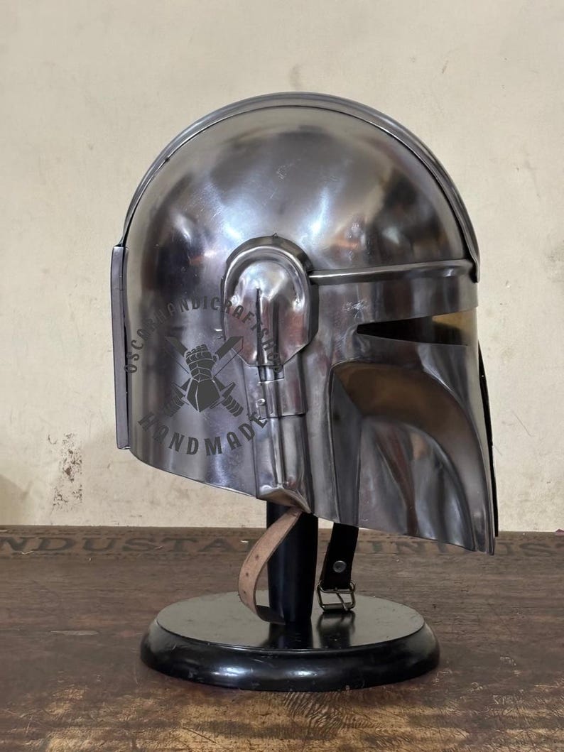 May include: A silver-colored helmet with a visor and ear pieces, displayed on a black stand. The helmet has a reflective surface and appears to be made of metal. The text "HANDMADE" is visible on the side.