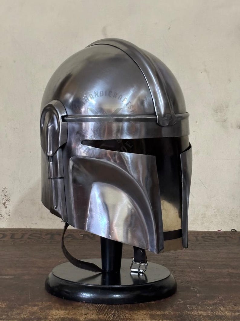 May include: A metallic silver Mandalorian helmet, displayed on a black stand. The helmet features a visor, ear pieces, and a smooth, reflective surface. The helmet is a replica of the one worn by the character in the Star Wars series.