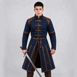May include: A long, quilted blue gambeson jacket featuring brown leather shoulder panels, trim, and vertical straps. The garment includes a high collar, a front closure with four brass-toned buckles, and a matching brown leather waist belt.