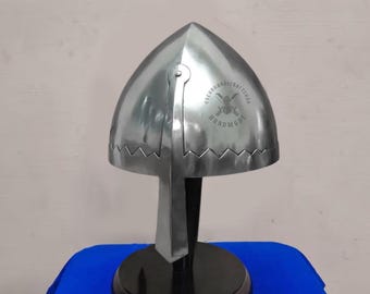 May include: A polished steel helmet with a pointed top, displayed on a black stand with a round base. The helmet has a decorative zigzag pattern along the bottom edge and a riveted hinge. The text "OSCARHANDICRAFT SHOP HANDMADE" is visible on the helmet.