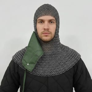 May include: A chainmail coif with a hood and neck protection. The coif is made of small, interlocking metal rings. A green fabric piece with text is attached to the coif. The person is wearing a black quilted garment.