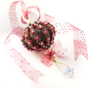 May include: A beaded Christmas bauble in shades of red, brown, and gold with a crystal drop hanging from a gold wire loop. The bauble is tied with a red ribbon with white polka dots.