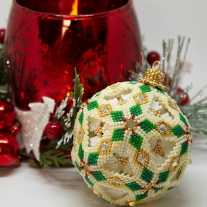 May include: A white and green beaded Christmas bauble with a gold trim, resting on a bed of green pine branches with red berries and a red glass candle holder.