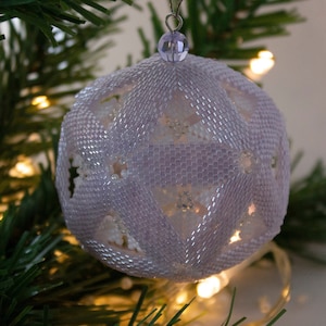 May include: A delicate, white, beaded Christmas ornament with a geometric design. The ornament is hanging from a green Christmas tree branch.