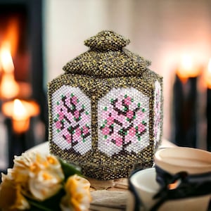 Könnte beinhalten: Eine sechseckige Perlenbox mit Deckel, die mit einem Blumendesign aus rosa und weißen Blüten auf braunem Hintergrund verziert ist.