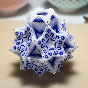 Pode incluir: Uma esfera de contas brancas e azuis com um design geométrico. A esfera é feita de contas pequenas e tem um padrão em forma de estrela.