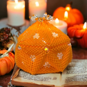 May include: A beaded orange pumpkin-shaped decorative object with a crystal-like top. The object is sitting on a book with a brown cover.