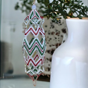 May include: A white, red and green beaded Christmas bauble with a geometric design. The bauble is shaped like a teardrop and has a silver loop at the top.