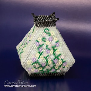 May include: A white beaded vase with a floral design in shades of purple, green, and yellow. The vase has a black metal rim and base.